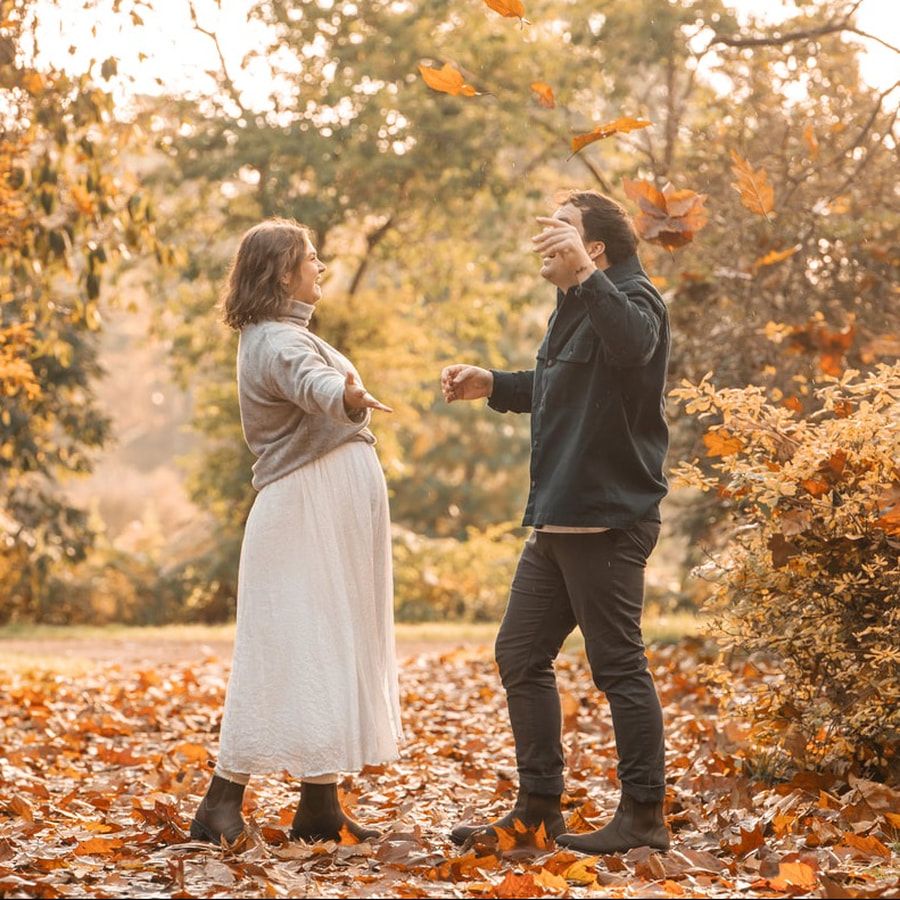 A couple walks through a park filled with fallen autumn leaves and golden foliage in the late afternoon light.