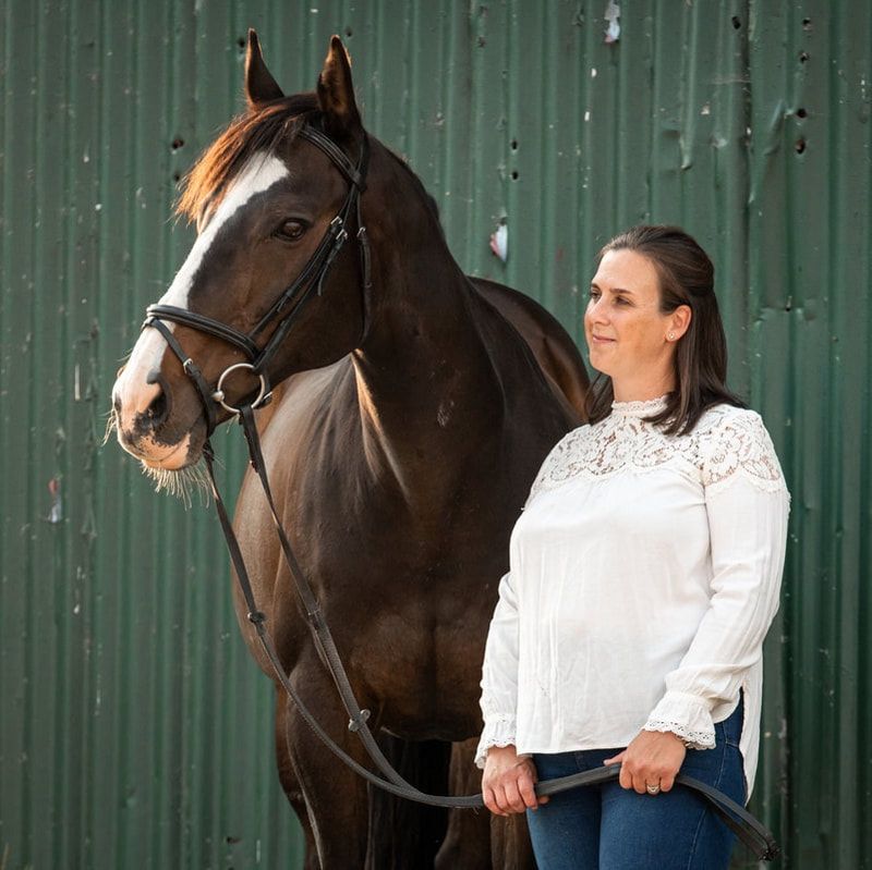 Woman in white sweater and jeans poses with dark brown horse against green metal wall in equestrian photoshoot.