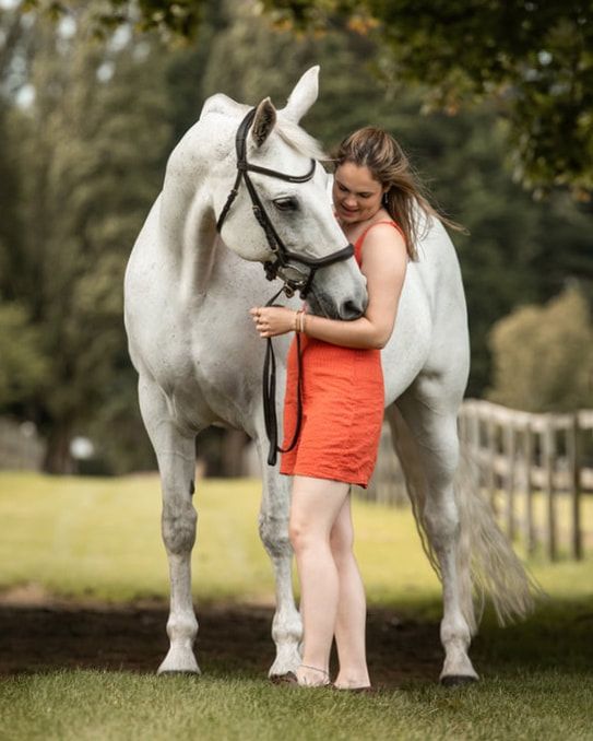 A white horse and someone in an orange dress share a tender moment together in a grassy field.