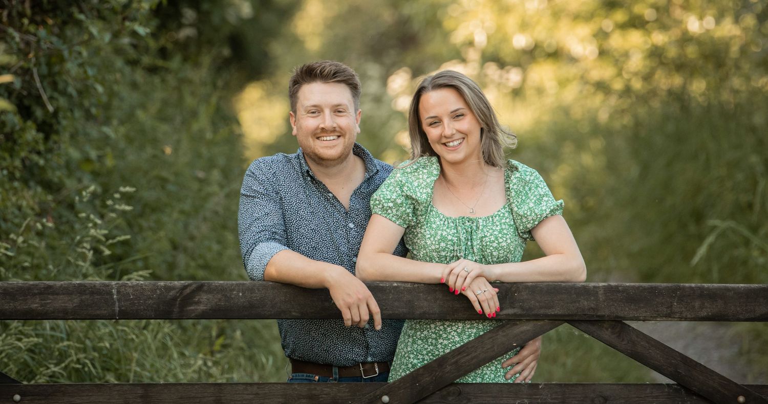 A couple sits together on a rustic wooden fence during a golden sunset in a natural outdoor setting.