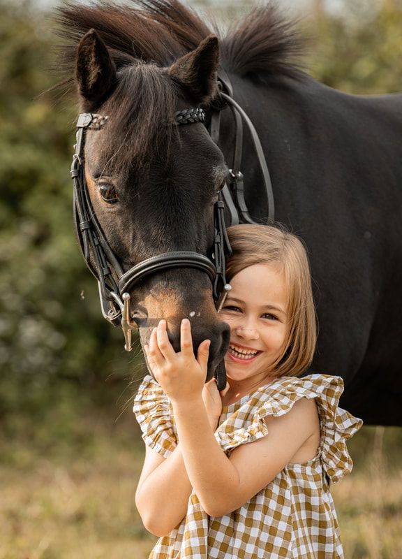 A young child in a gingham dress shares a tender moment with a black horse in a sunlit outdoor setting.