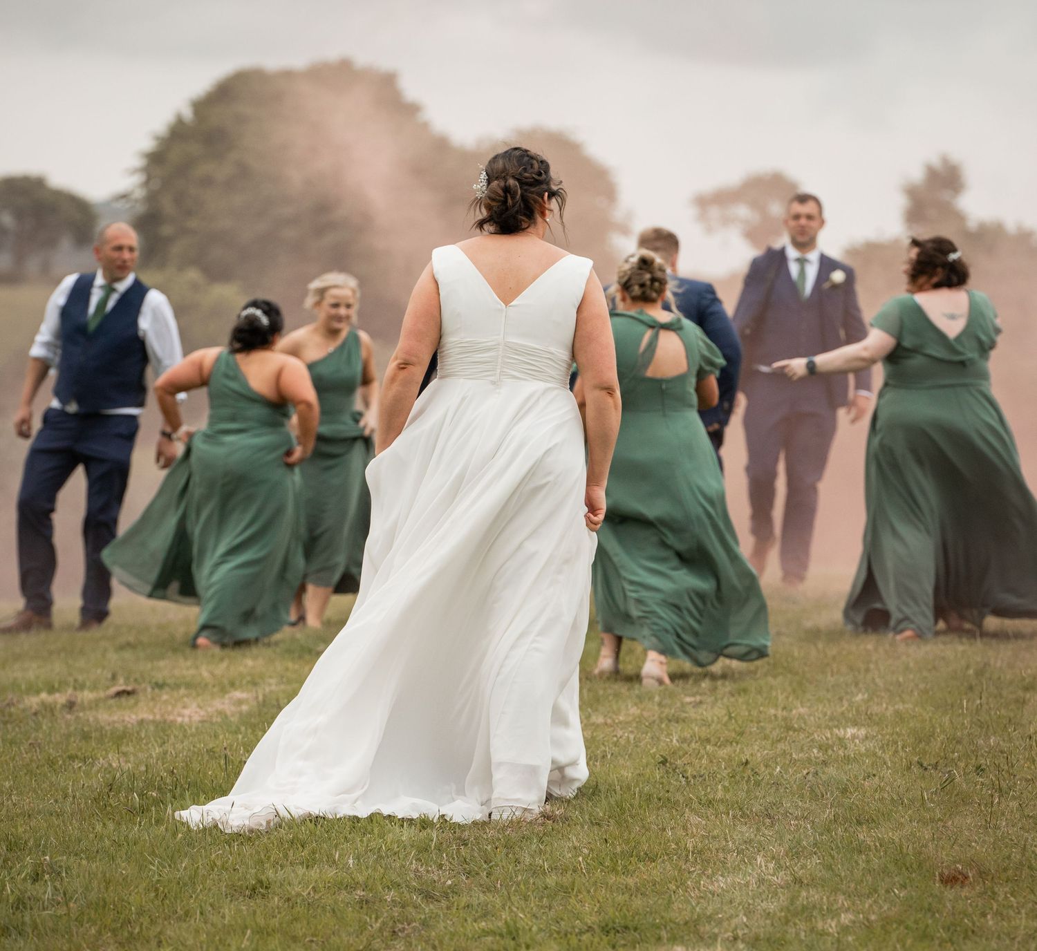 Wedding party in sage green dresses and suits poses playfully in a misty field at sunset.
