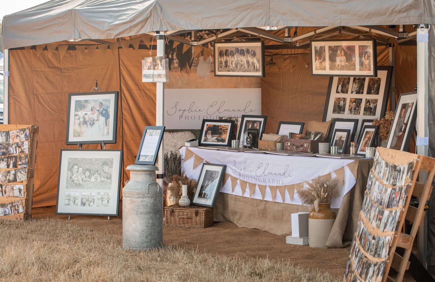 Rustic outdoor vendor booth displaying framed artwork and family portraits with burlap decor at a craft fair.