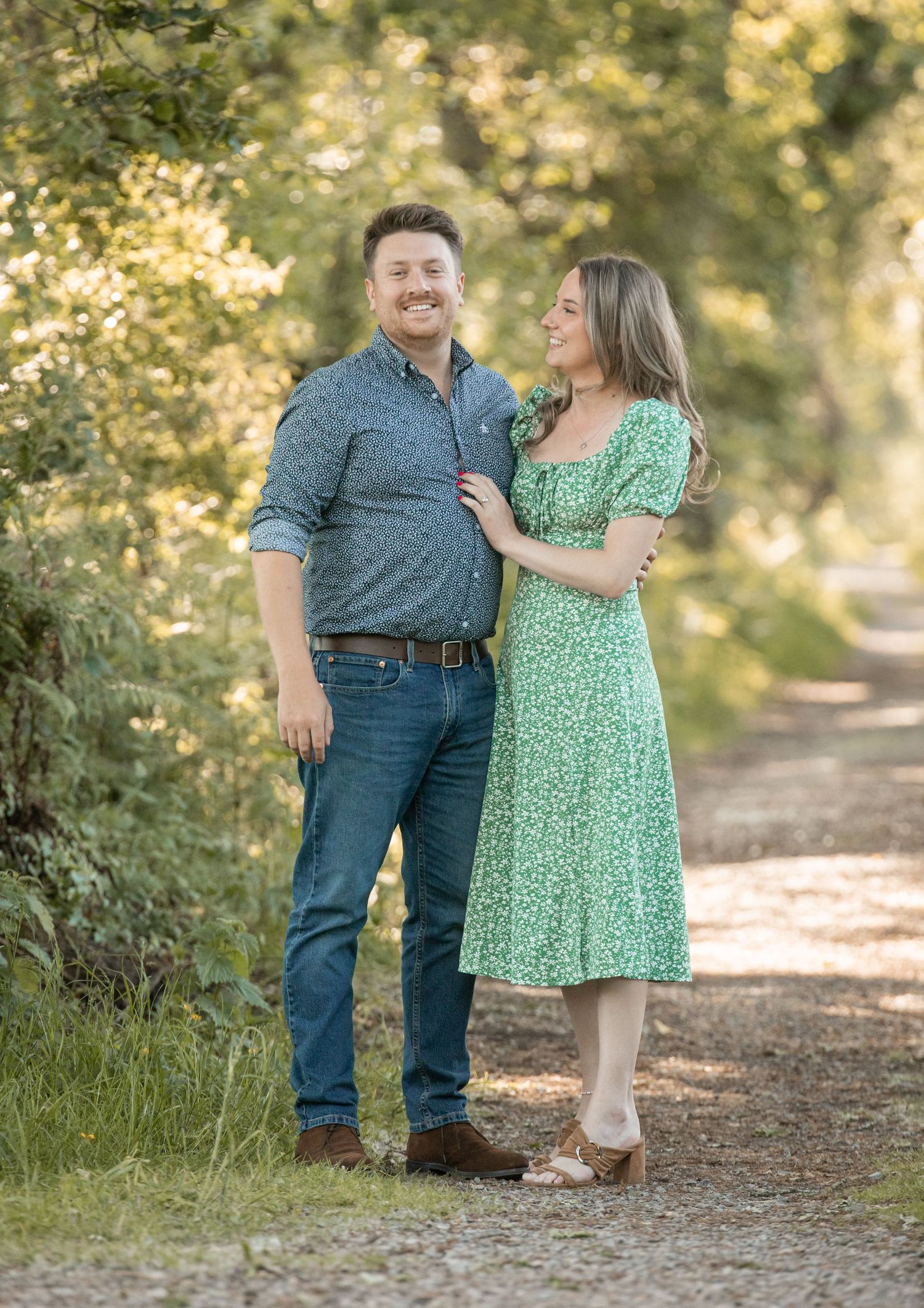 A couple shares a tender moment on a sunlit path surrounded by lush greenery and trees.