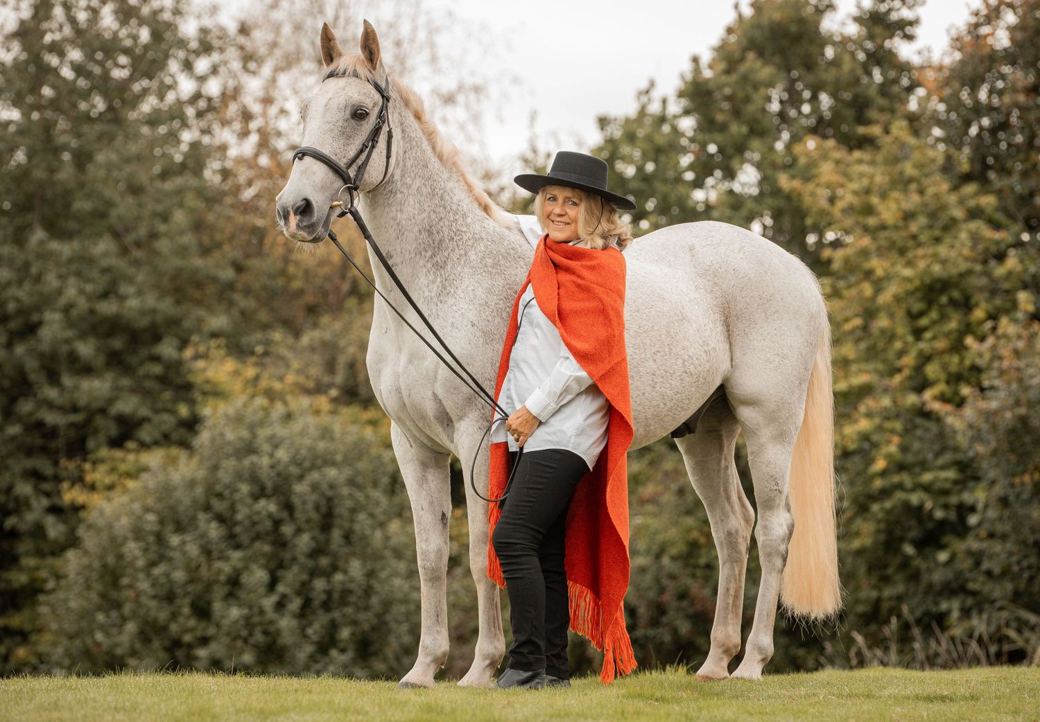 A white horse stands gracefully in a lush green field wearing a bright orange saddle blanket.