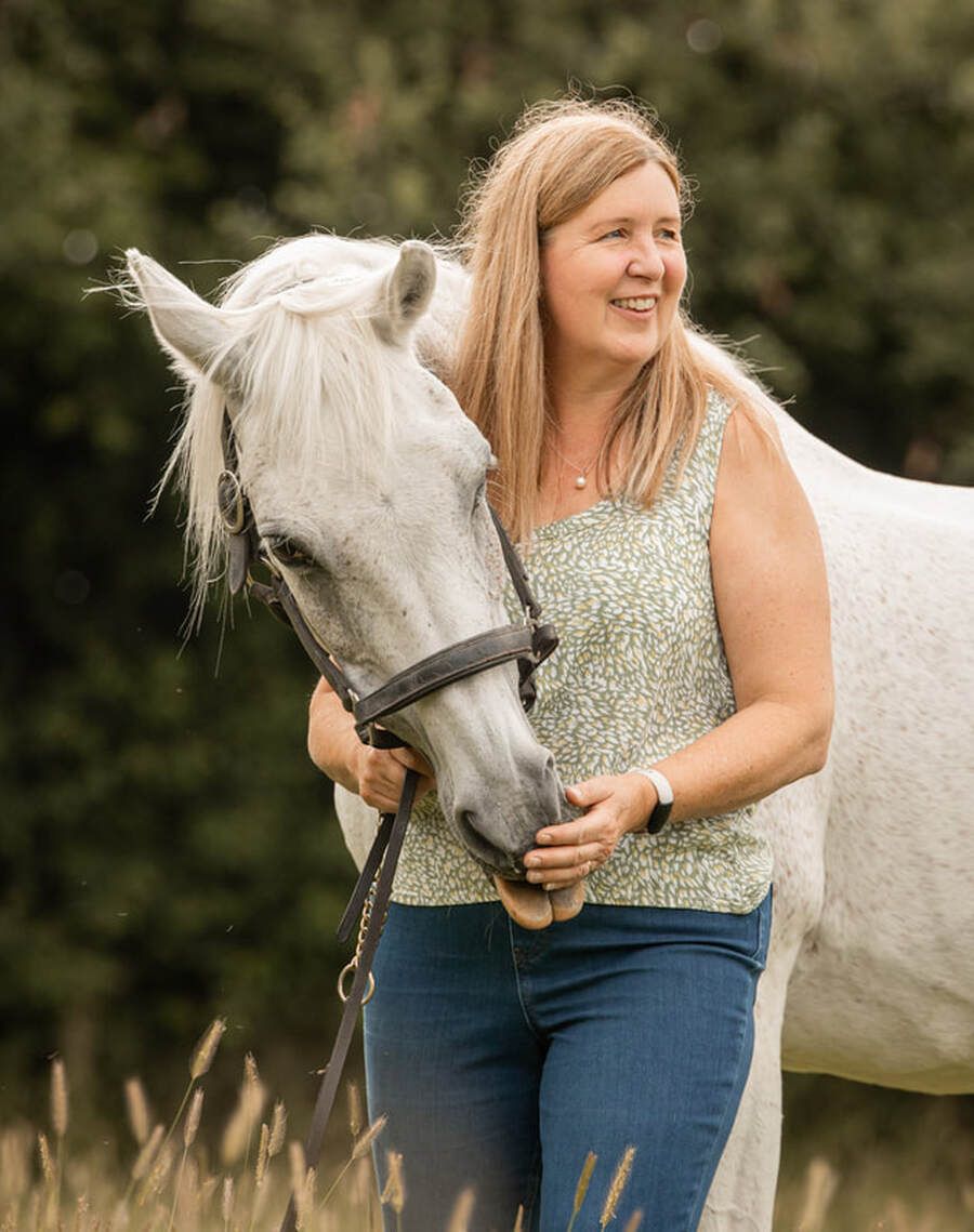 A woman in a floral top and jeans interacts affectionately with a white horse in a natural outdoor setting.