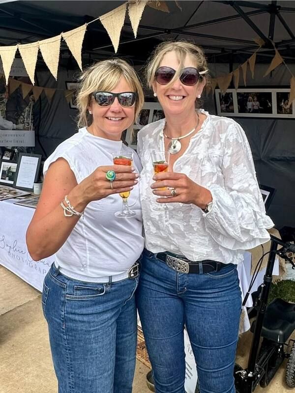 Two friends in white tops and jeans hold up colorful drinks while celebrating at an outdoor event under bunting.