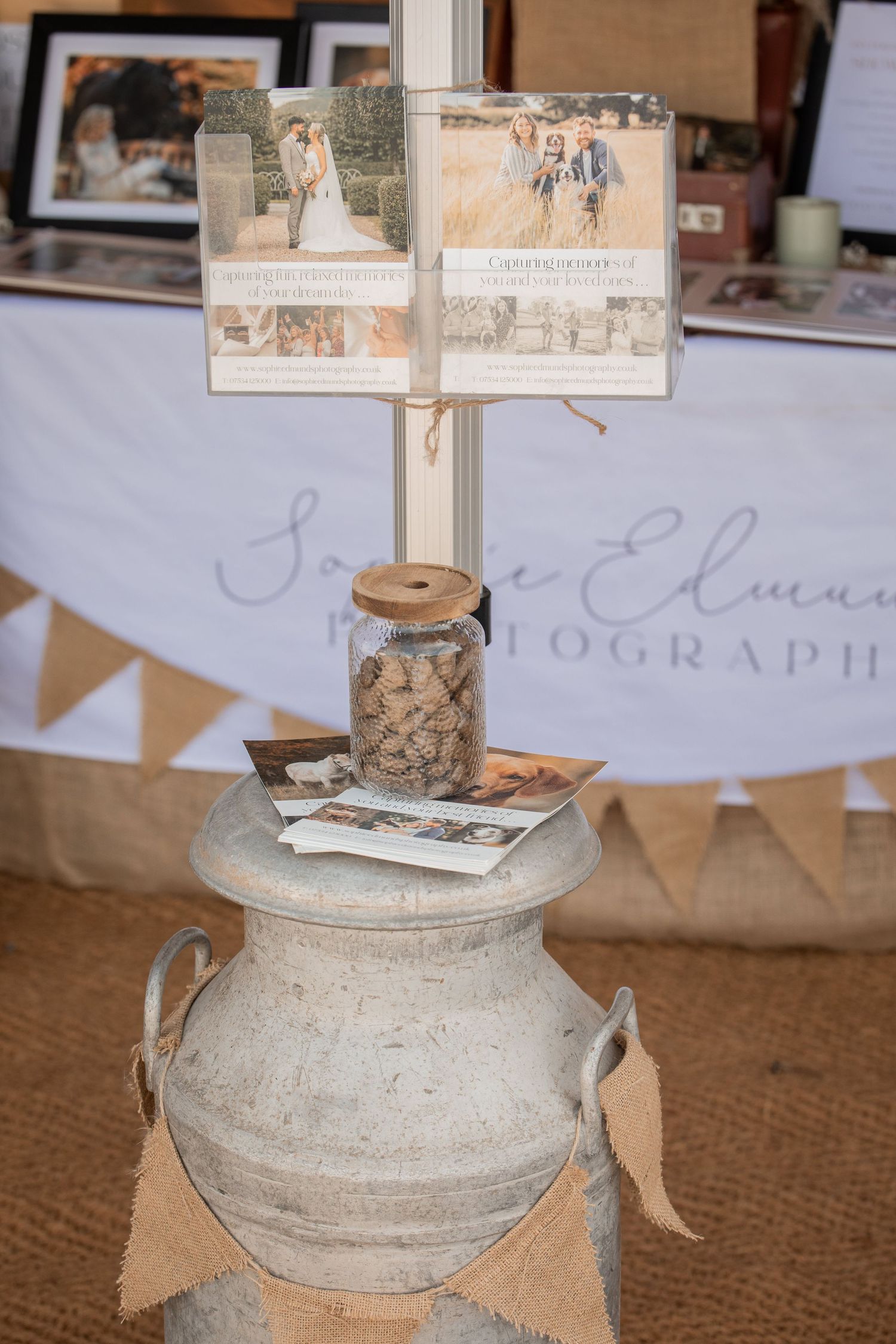Rustic wedding photography display on vintage milk can with burlap bunting, mason jar, and photo samples at a bridal fair.