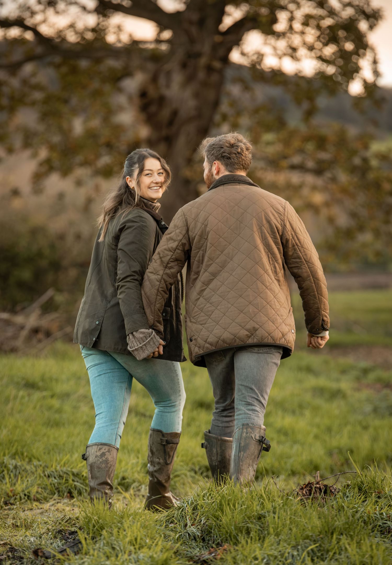 A couple holding hands while walking through a grassy field at sunset wearing casual outdoor clothing and boots.