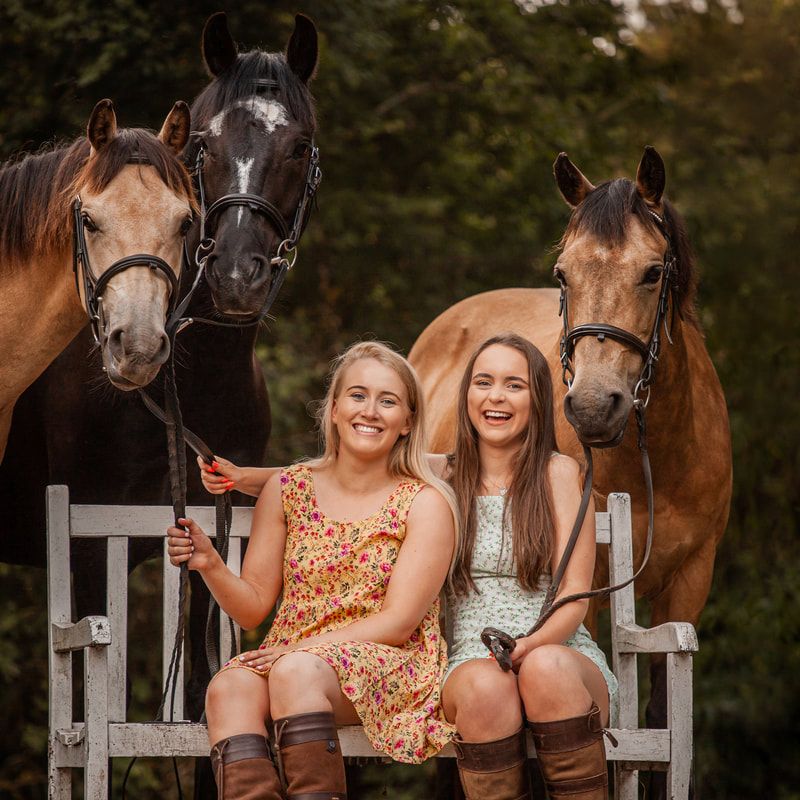 Two people share laughs while posing with horses at a rustic fence.