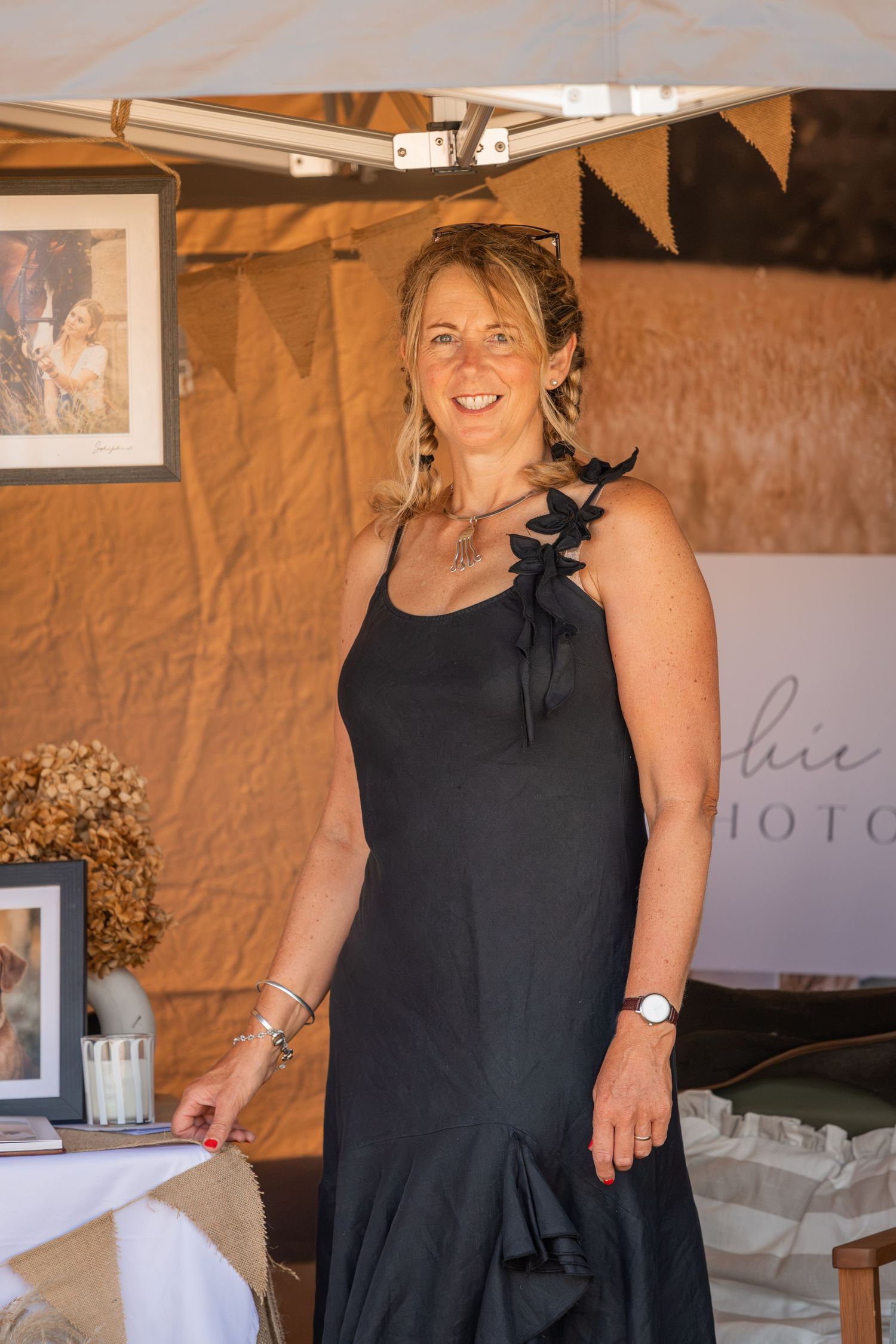 Smiling blonde woman in black dress standing at an outdoor photography vendor booth display.