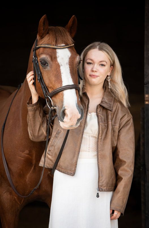 A woman in a brown leather jacket and white dress poses with a chestnut horse wearing a decorative bridle.