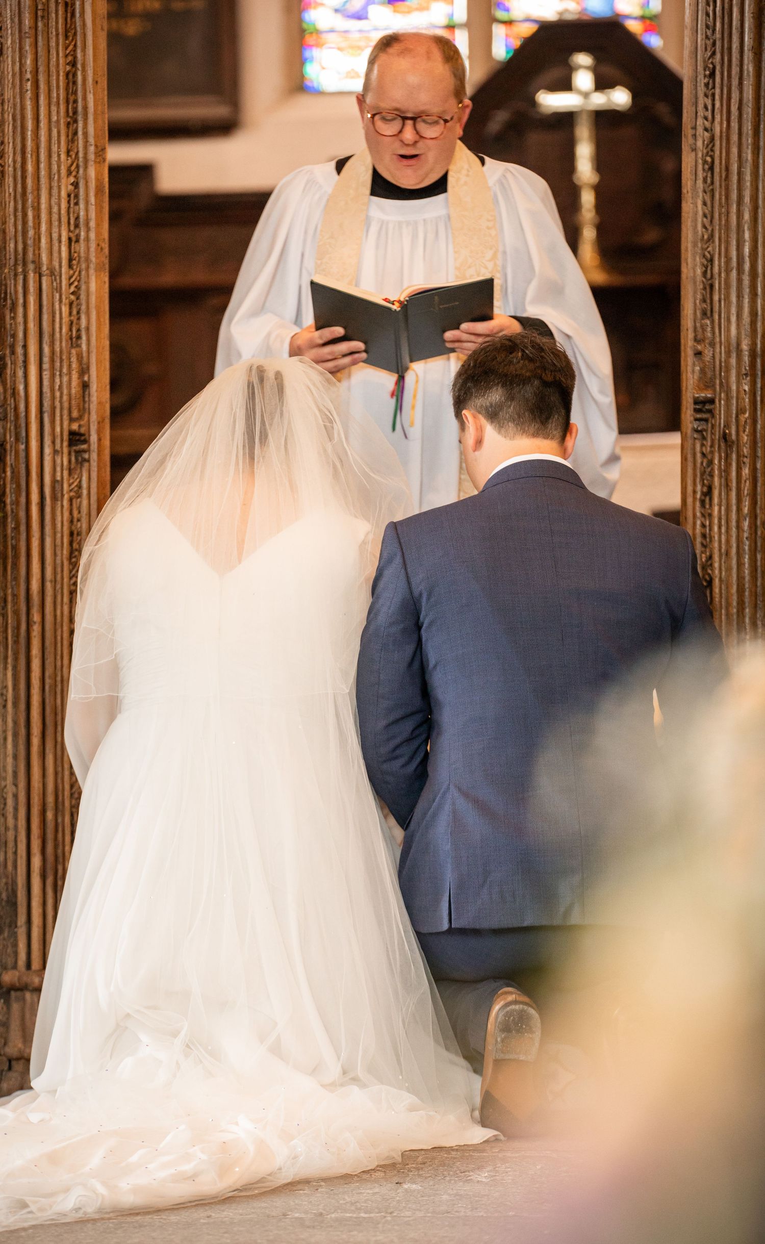 A wedding ceremony takes place in a church with stained glass windows as the couple stands before a minister.