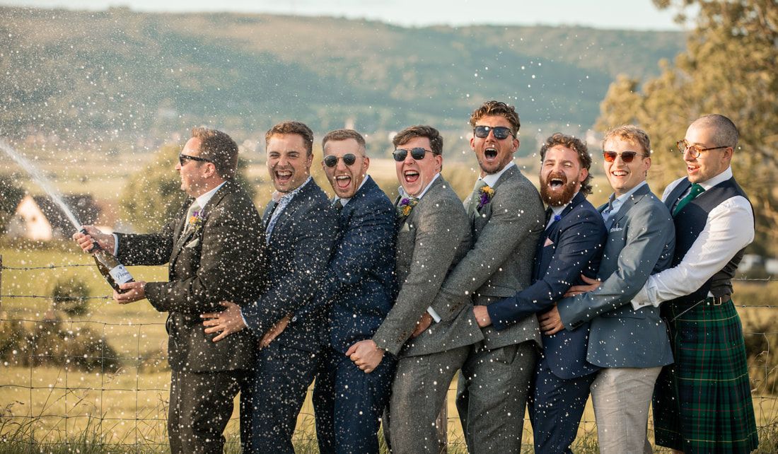 Groomsmen in formal attire stand in a line outdoors against a scenic autumn mountain backdrop.