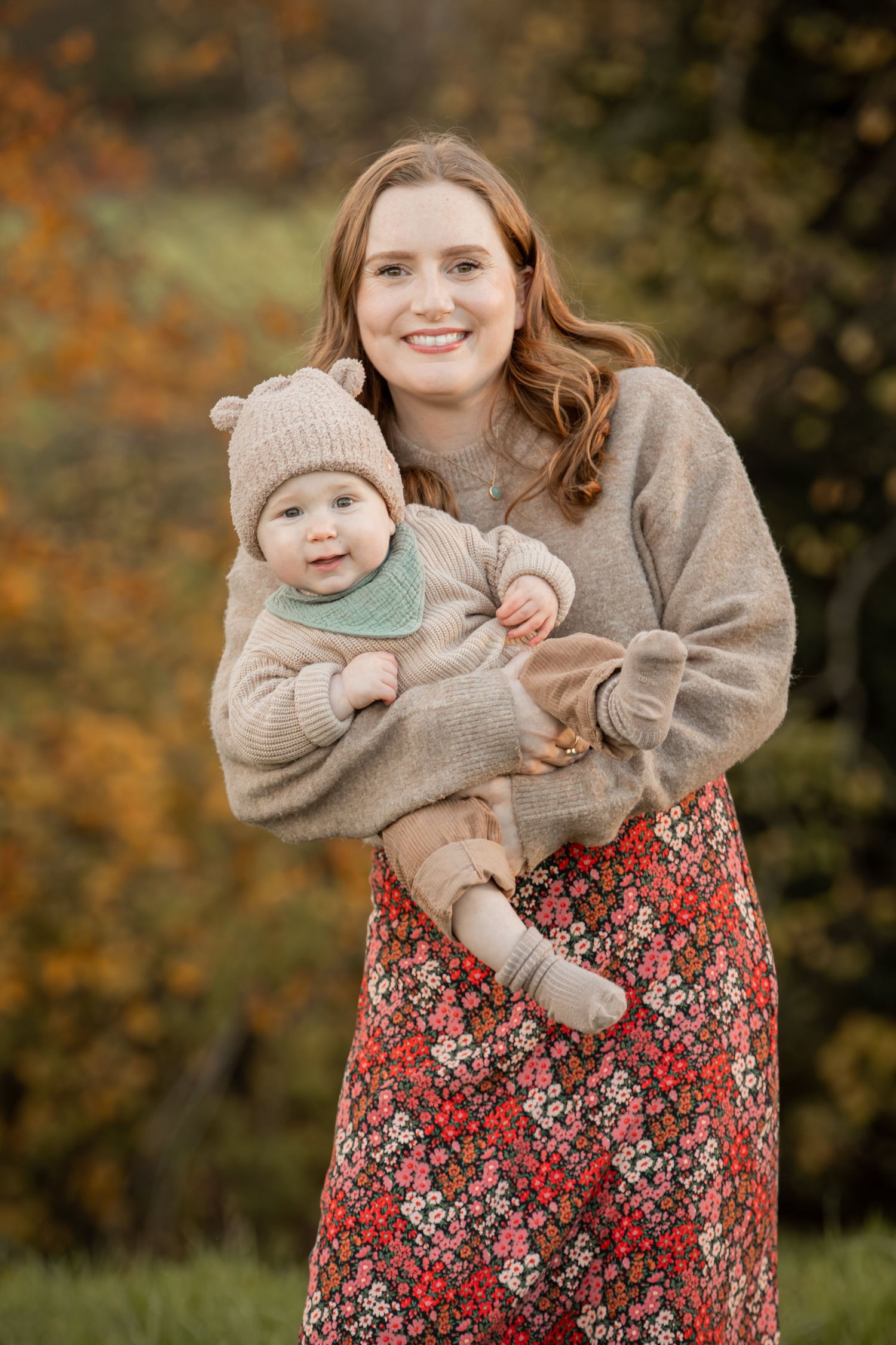 A mother in a beige sweater and floral skirt holds a baby wearing a neutral-colored outfit in an autumn outdoor setting.