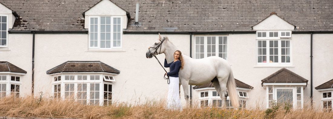 A panoramic view of a white coastal house with horses grazing in the foreground against a beige grassy landscape.