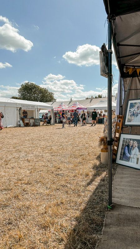 Outdoor market stalls and vendor tents set up along a dirt path on a sunny day with white puffy clouds in the sky.