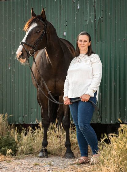 A dark brown horse stands beside someone in a white lace top and jeans against a green metal wall background.