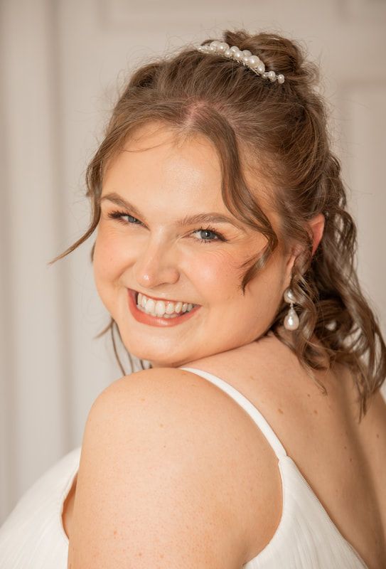 A bride wearing a pearl headpiece and white dress strap smiles warmly over her shoulder in a soft-lit portrait.