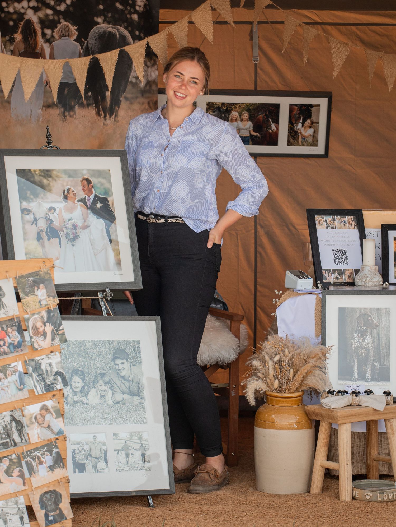 Woman standing in art display booth with framed portrait paintings and wedding photos at an outdoor market event.