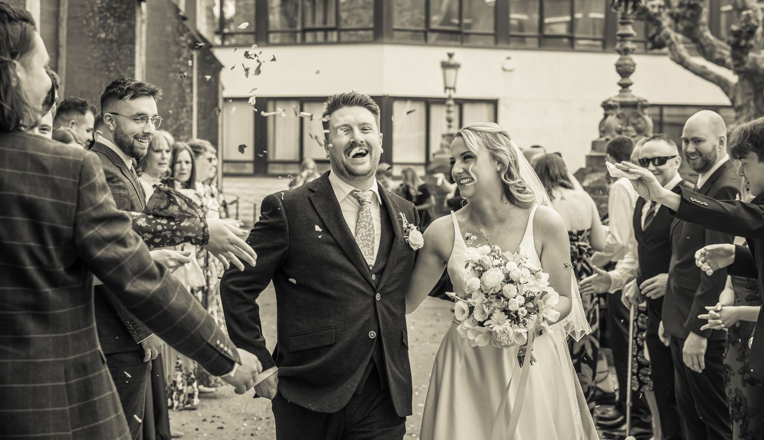 Black and white photo of a joyful bride and groom walking through confetti after their wedding ceremony.