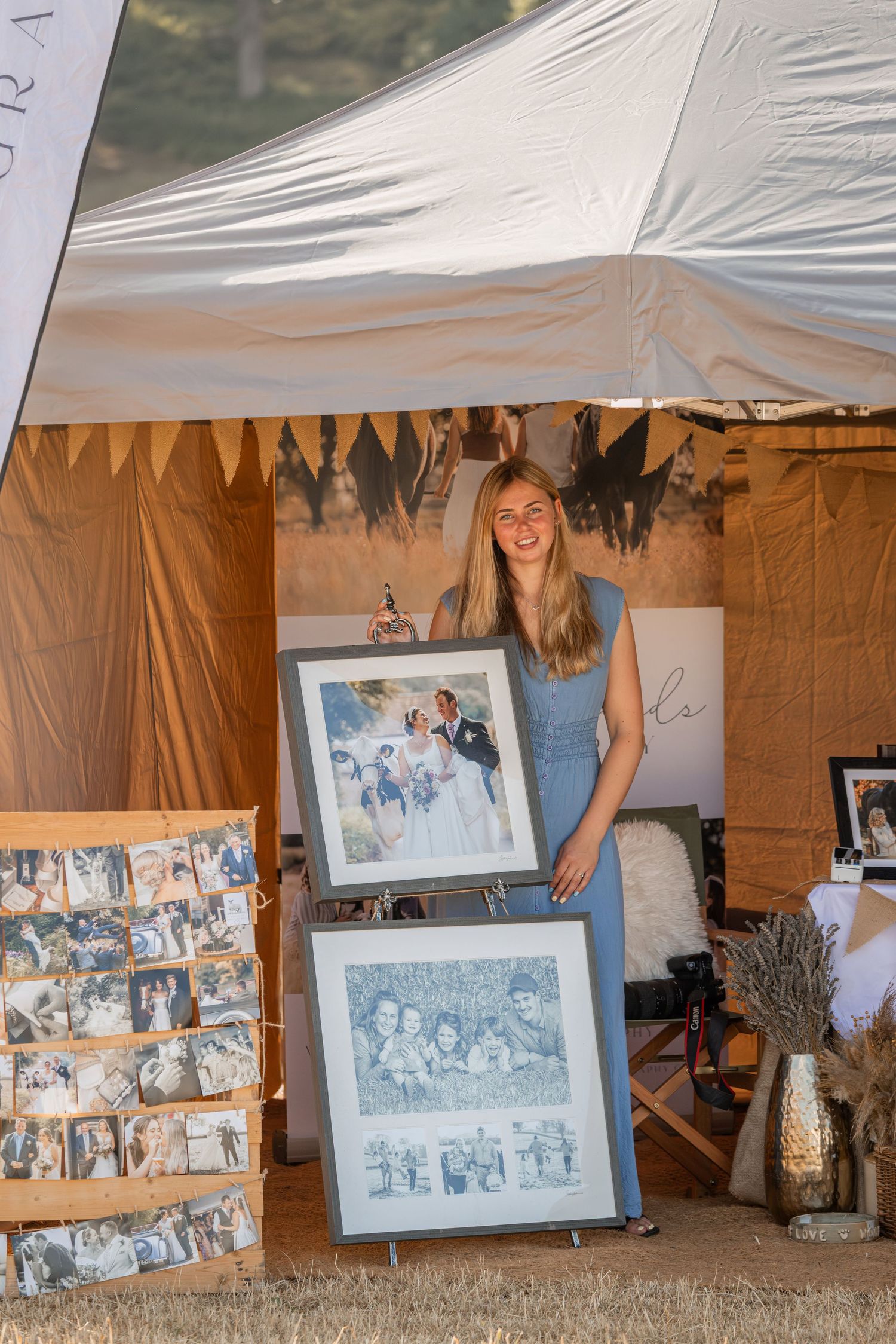 Woman in blue dress holding framed portrait artwork at an outdoor vendor booth with photo displays.