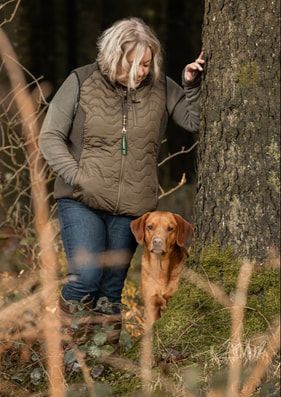 A Vizsla dog stands in tall grass next to someone wearing jeans and a brown jacket on an autumn day.