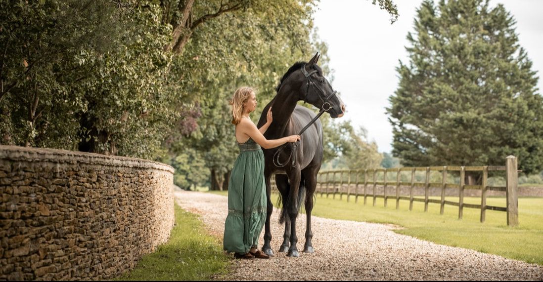 A person in an elegant green dress stands beside a majestic black horse on a gravel path near a stone wall.