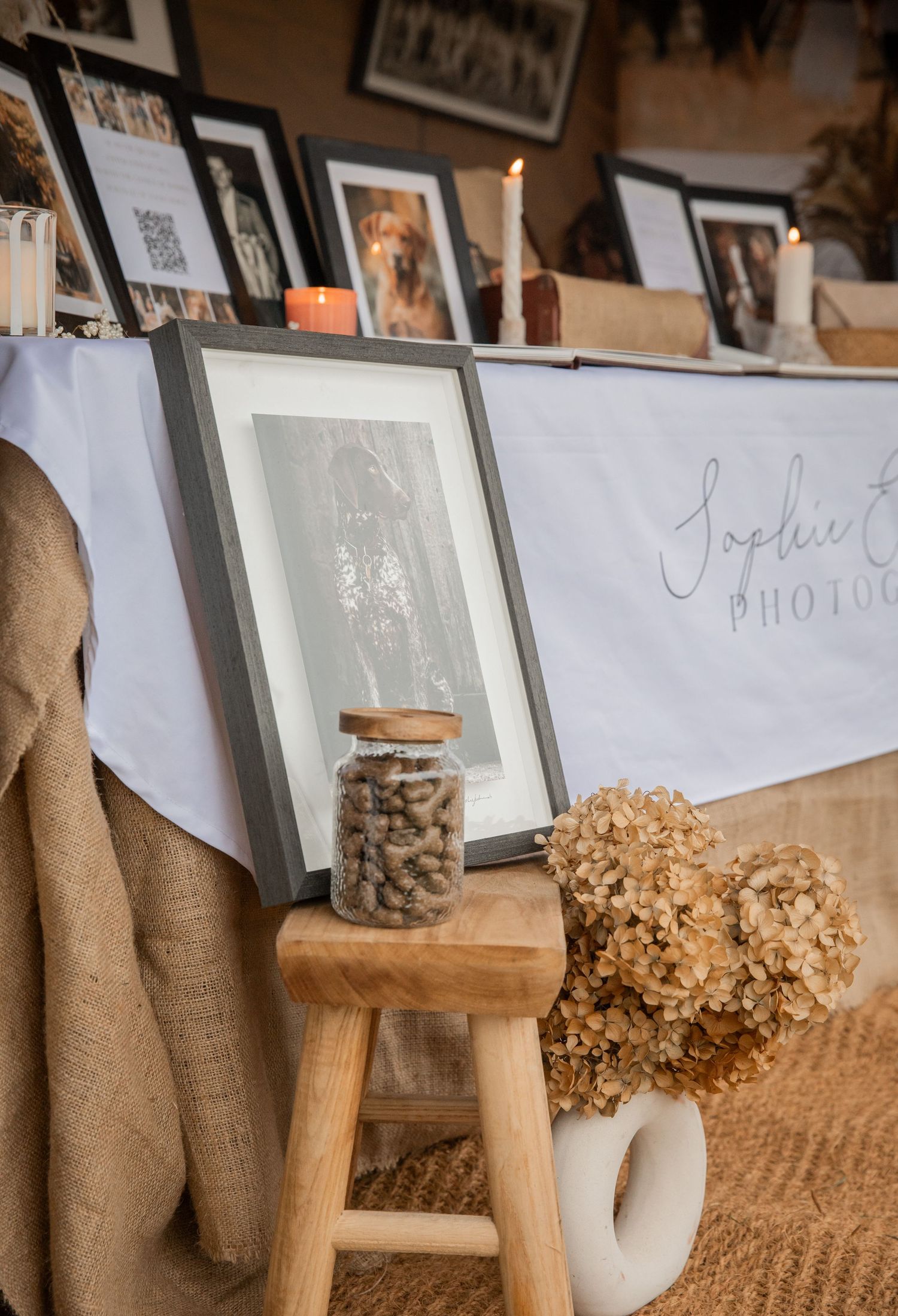 Rustic wedding display with framed photos, candles, dried hydrangeas, and a jar of nuts on a wooden stool.