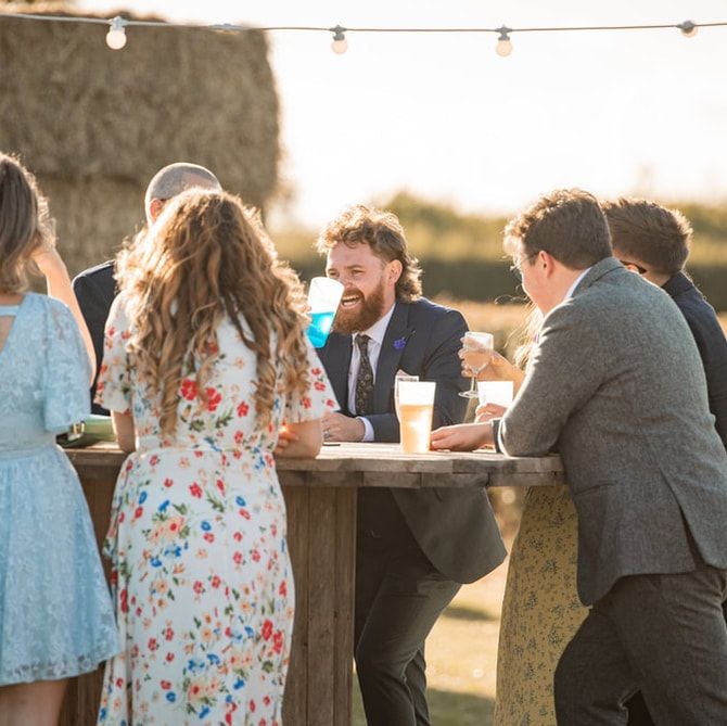 Outdoor wedding reception scene with string lights and guests gathered at rustic wooden tables during golden hour.