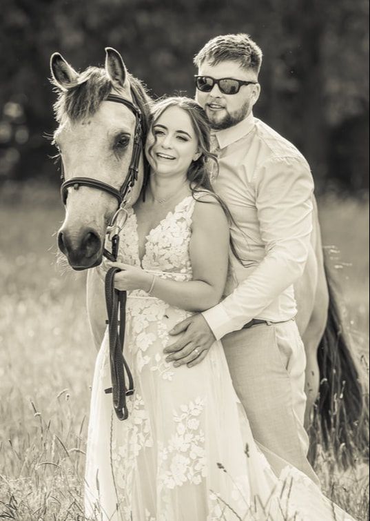 A couple in wedding attire poses with a white horse in a sunny meadow, captured in black and white photography.