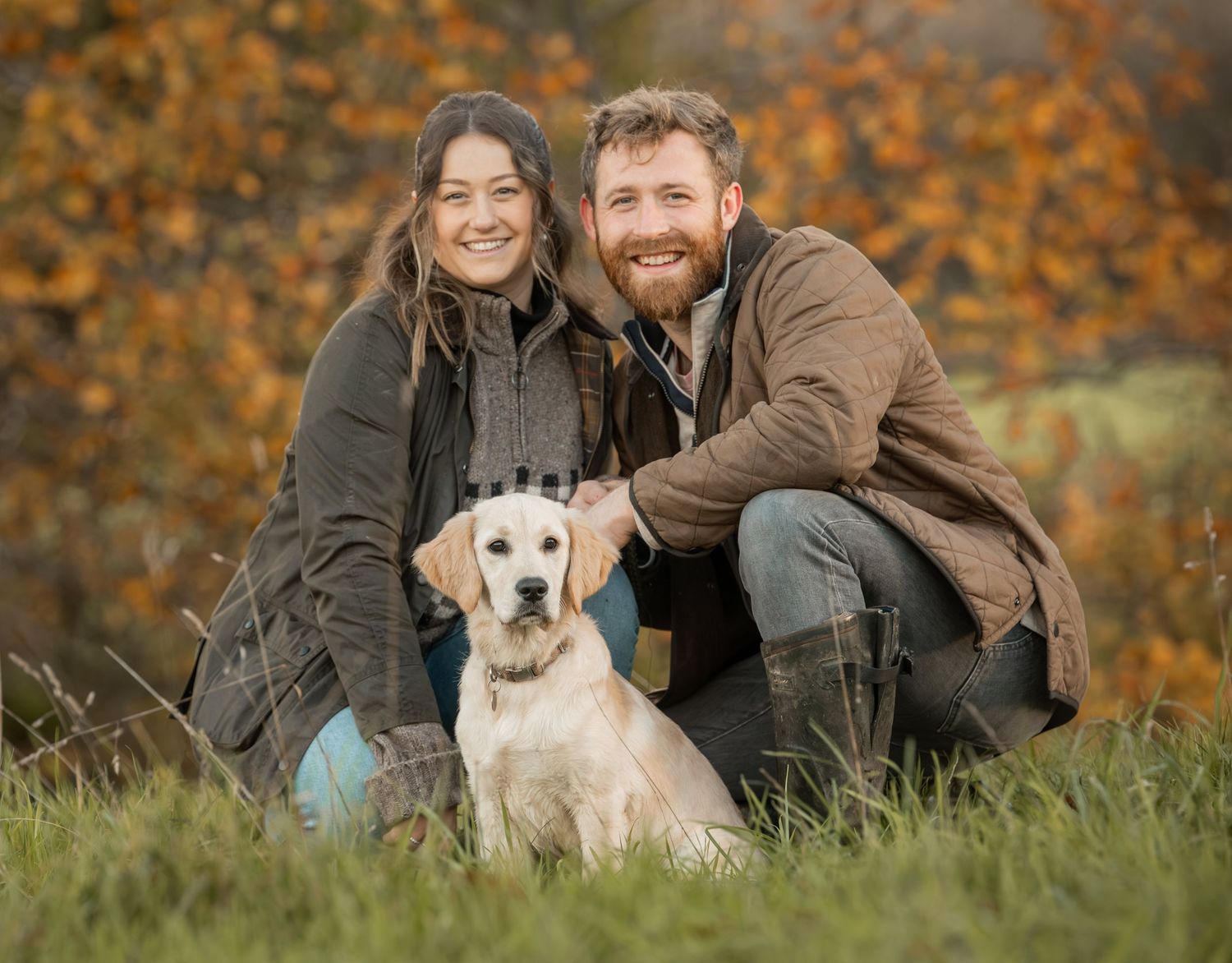 A couple sits with their white dog on grass surrounded by vibrant autumn foliage in the background.