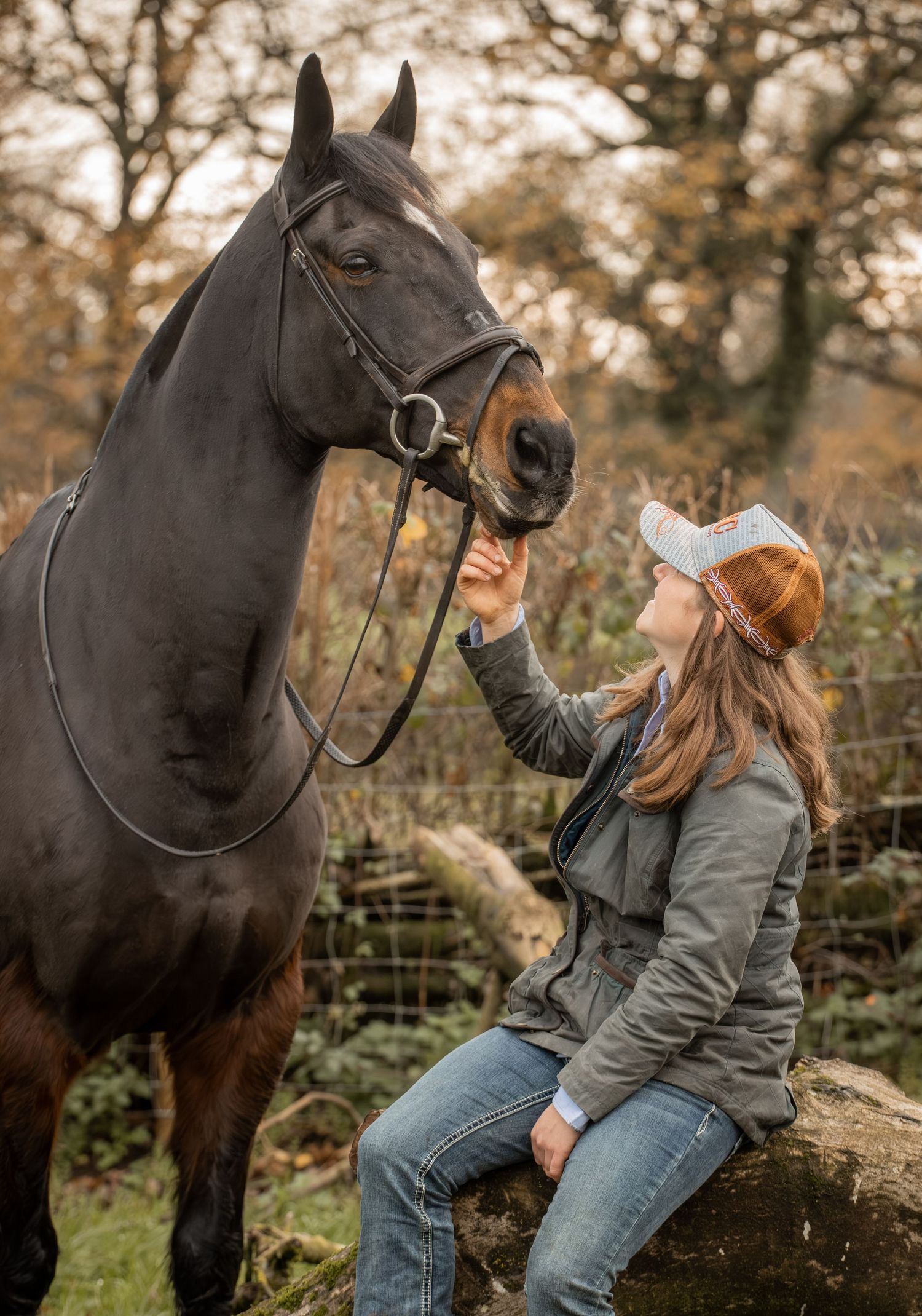 A dark brown horse and someone in a baseball cap share a tender moment in an autumn outdoor setting.