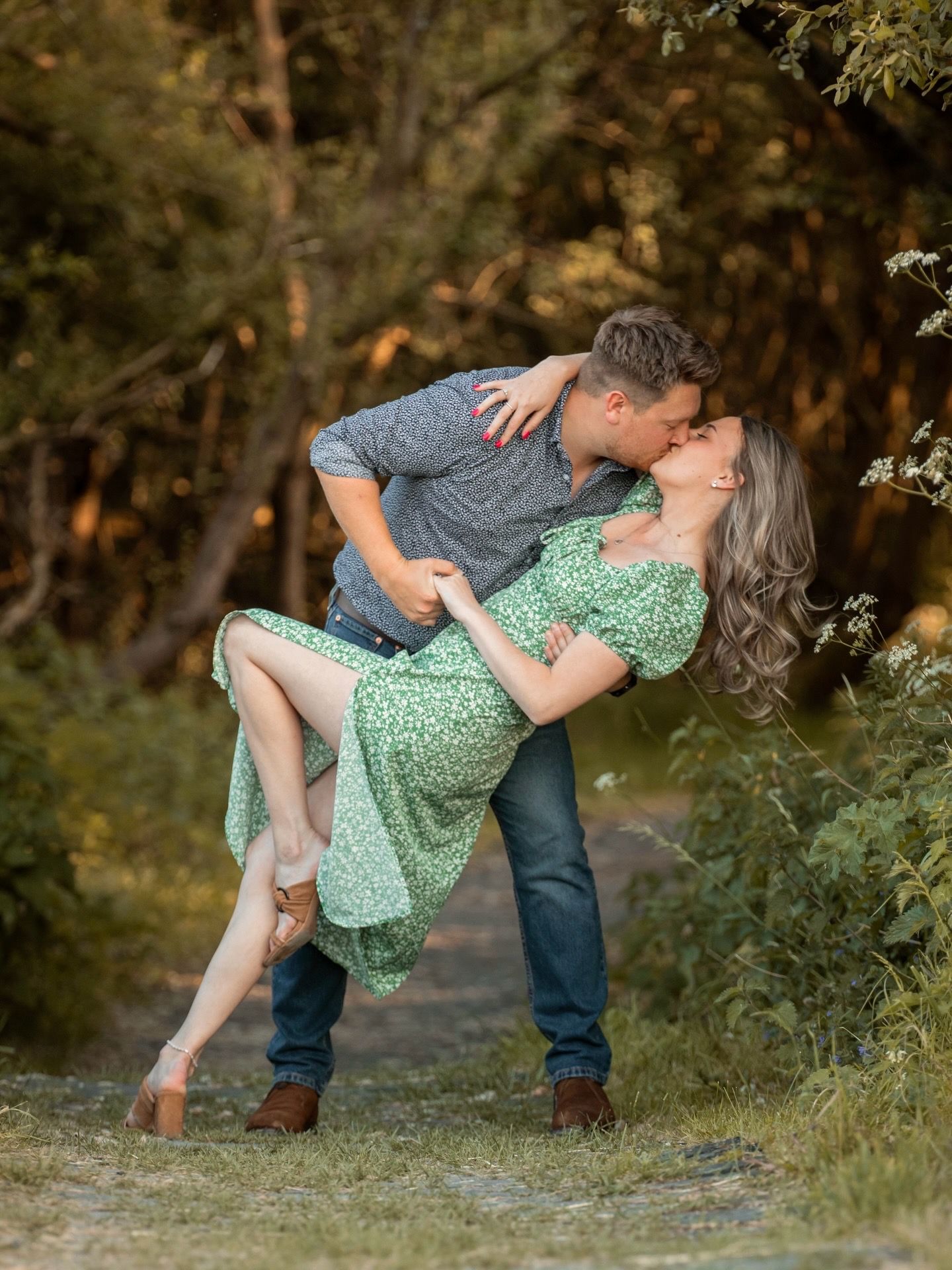 A romantic couple dances together in a dip pose outdoors during golden hour in a wooded setting.