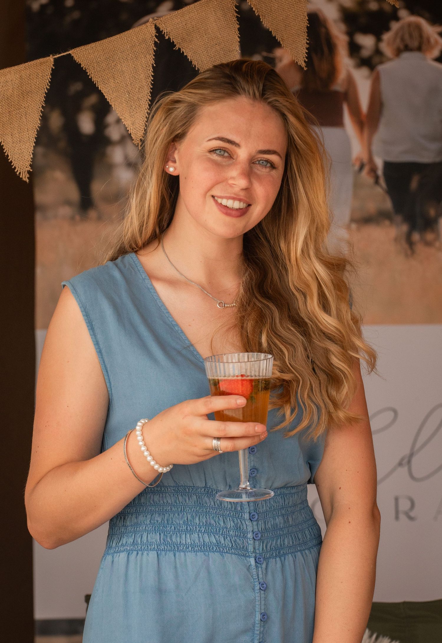 Young woman with long blonde hair smiling, holding a glass of wine at an outdoor event in a blue dress.
