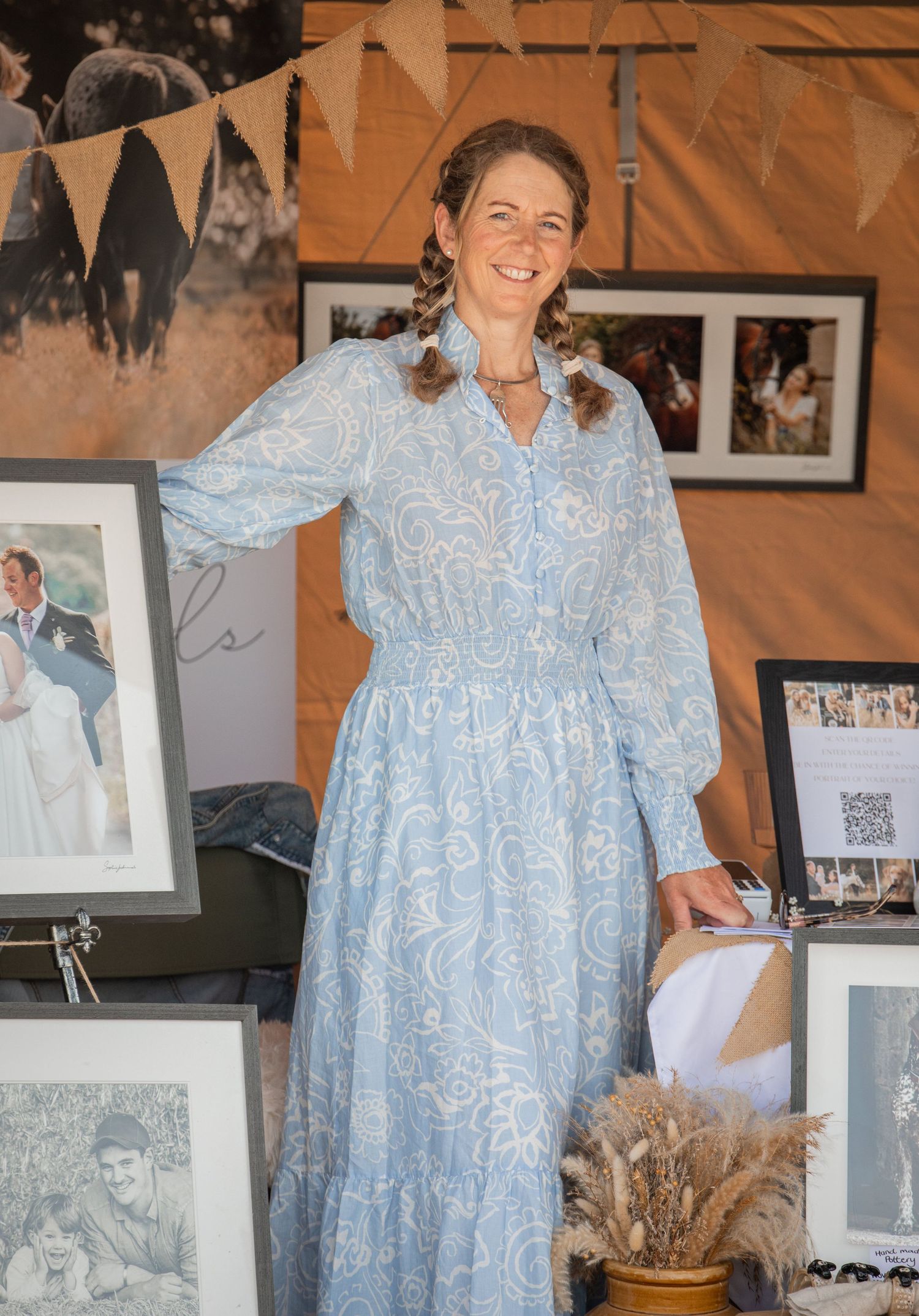 Woman in blue floral maxi dress smiling at an outdoor market stall displaying framed photographs.