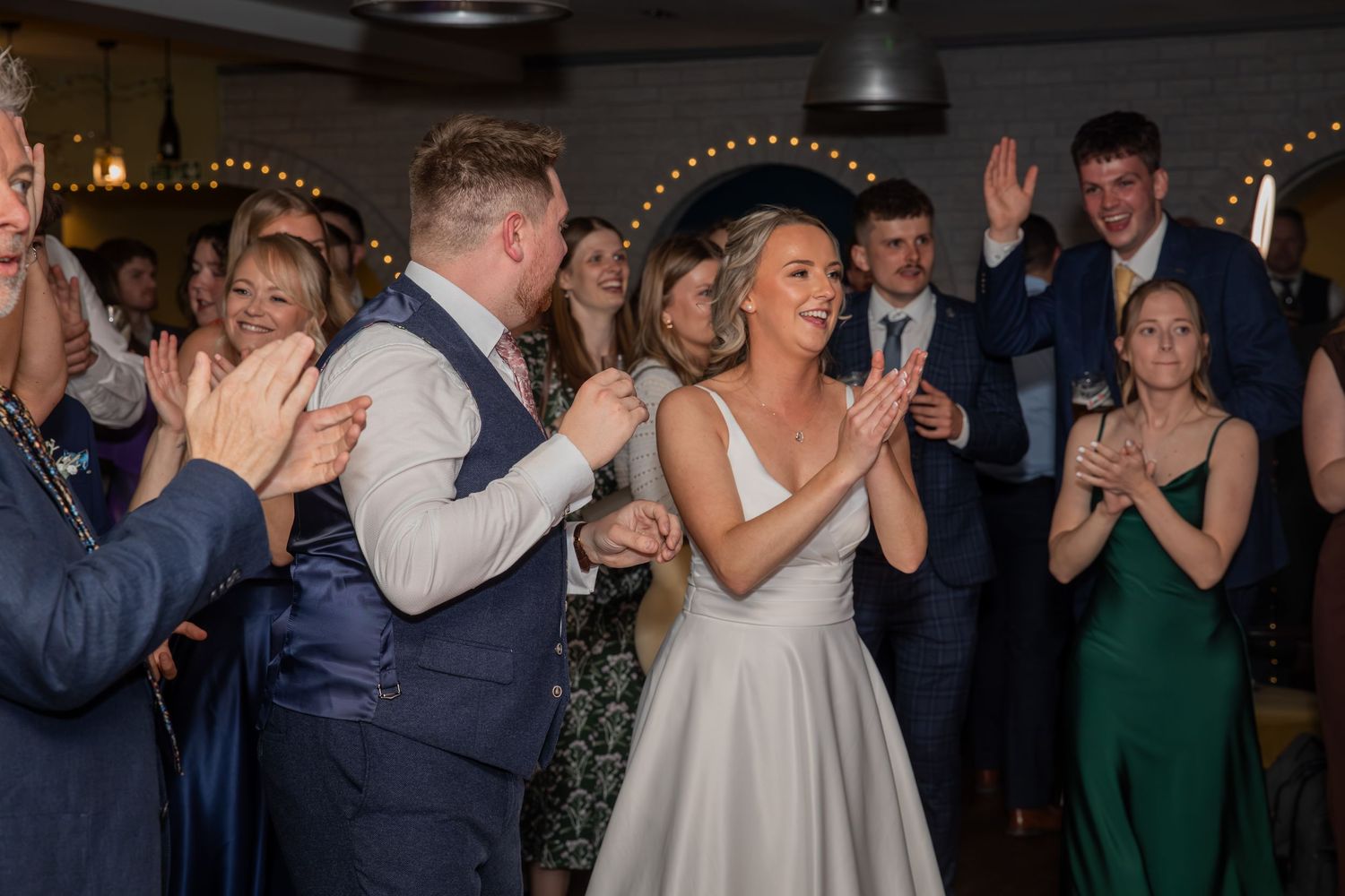 Bride and groom dancing joyfully with wedding guests clapping and celebrating at a stylish indoor wedding reception.