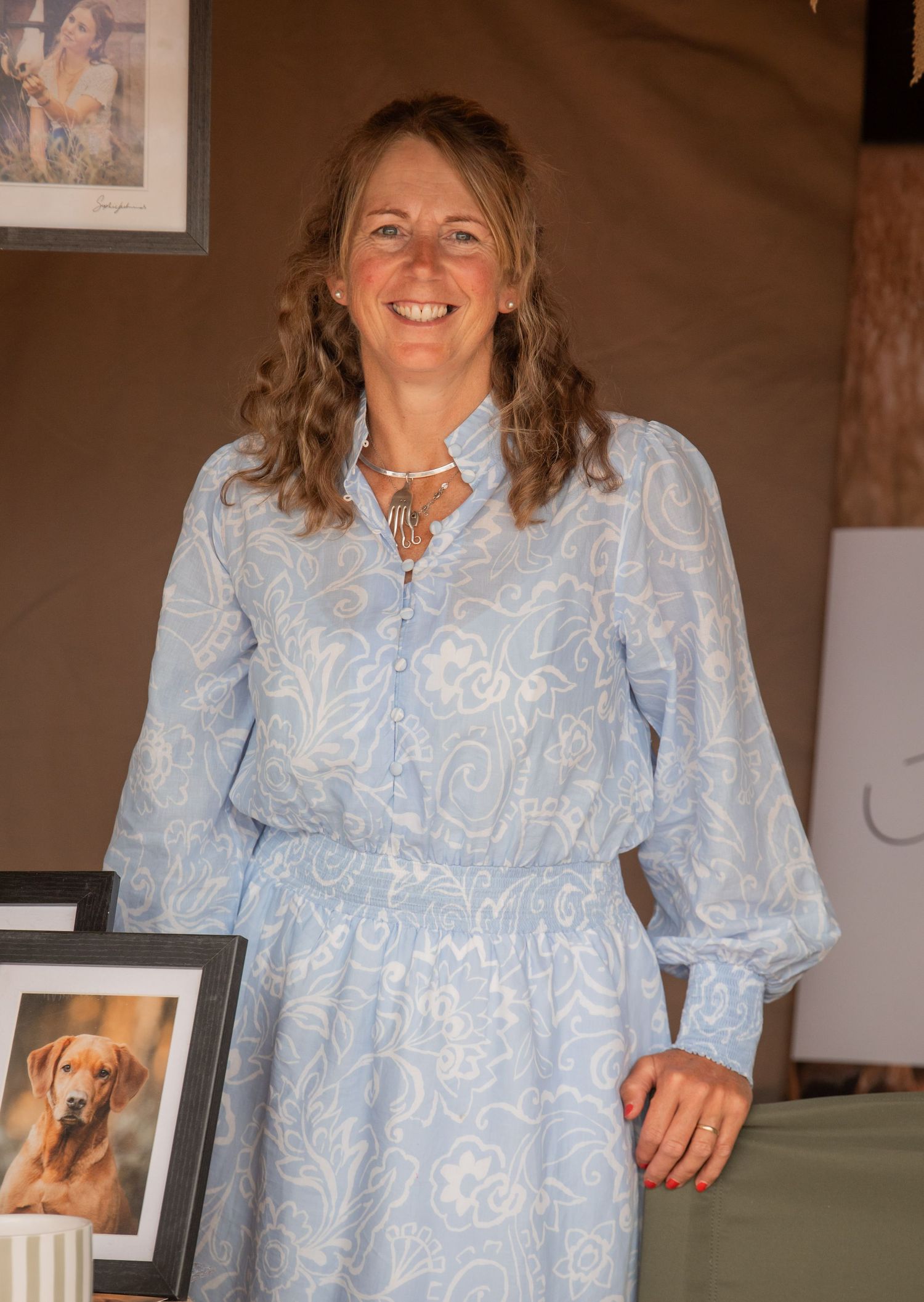 A smiling woman in a light blue paisley dress stands near framed dog and portrait artwork at an outdoor event.