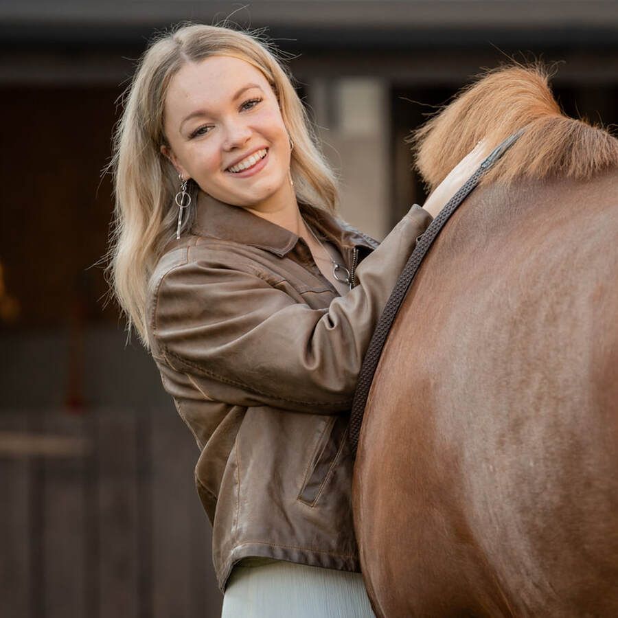 A person in a brown leather jacket smiles while leaning against a chestnut-colored horse at a stable.