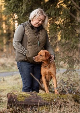 A person in a vest and jeans kneels beside a reddish-brown dog in an outdoor autumn setting with evergreen trees.