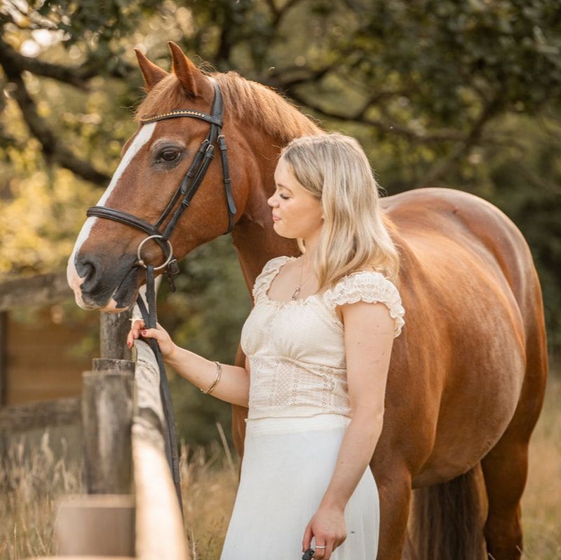 A woman in a white dress stands beside a chestnut horse in an outdoor setting during a photo session.