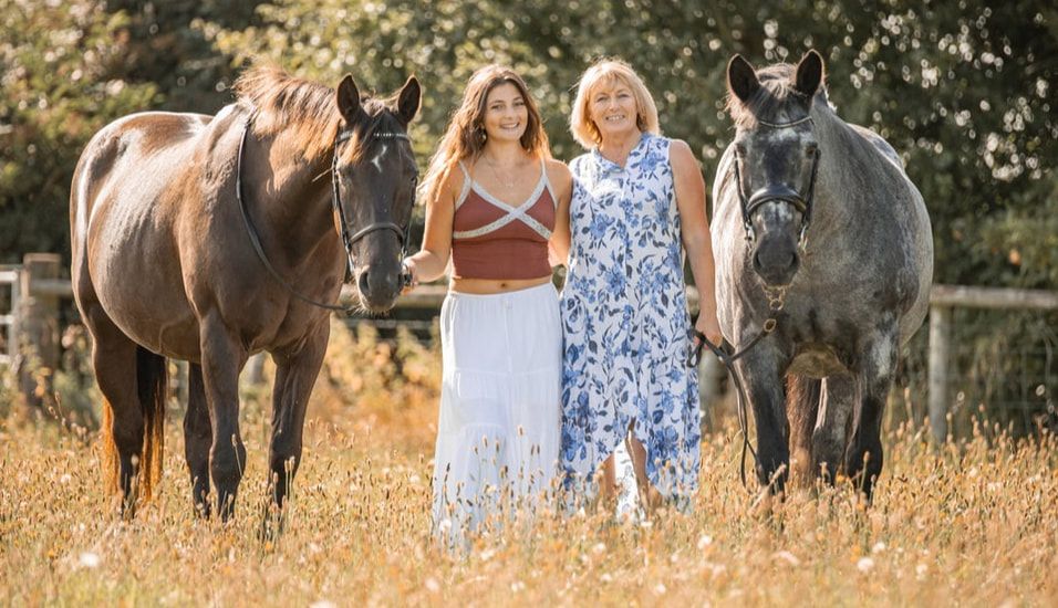 Two people in blue and white dresses walk through a field with horses grazing in the golden afternoon sunlight.