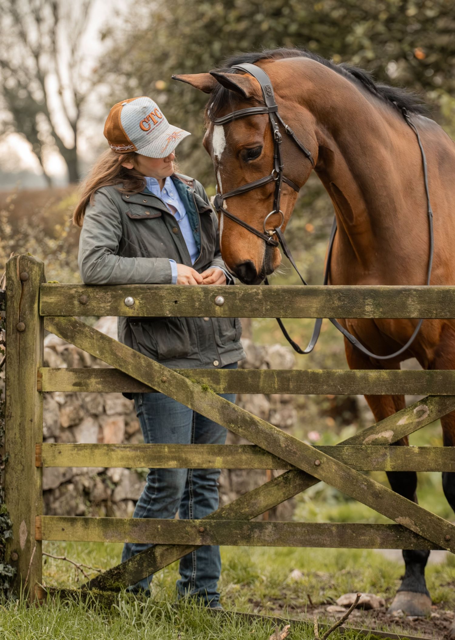 A person in a gray jacket stands by a wooden fence while interacting with a brown horse on a misty day.