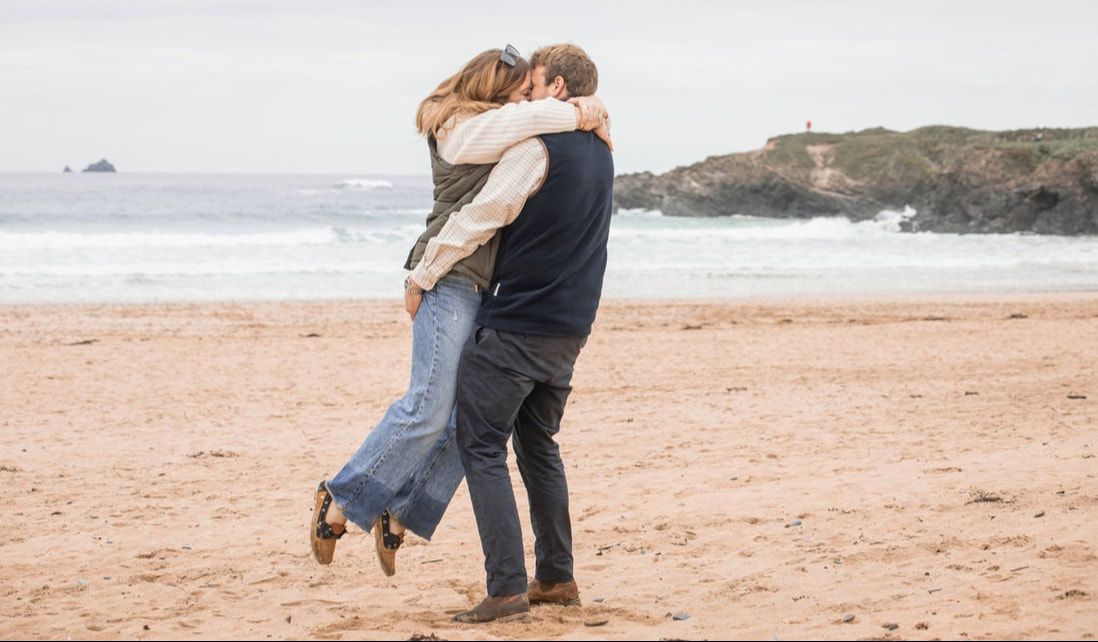 Romantic couple embracing on sandy beach with ocean waves and rocky coastline in background.