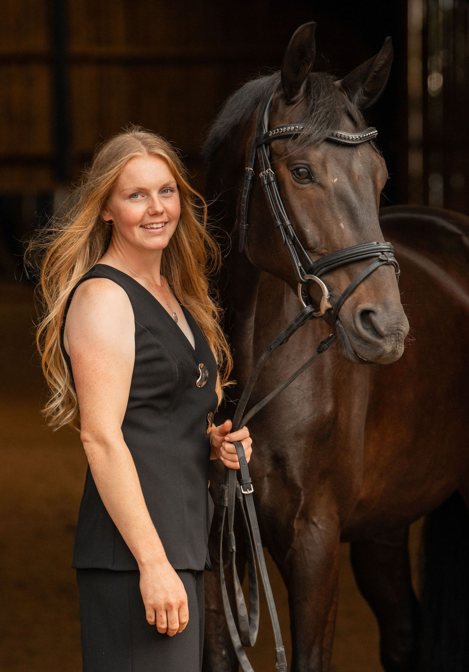 Caitlin Burgess Dressage Rider in black dress posing with dark horse in stable setting.