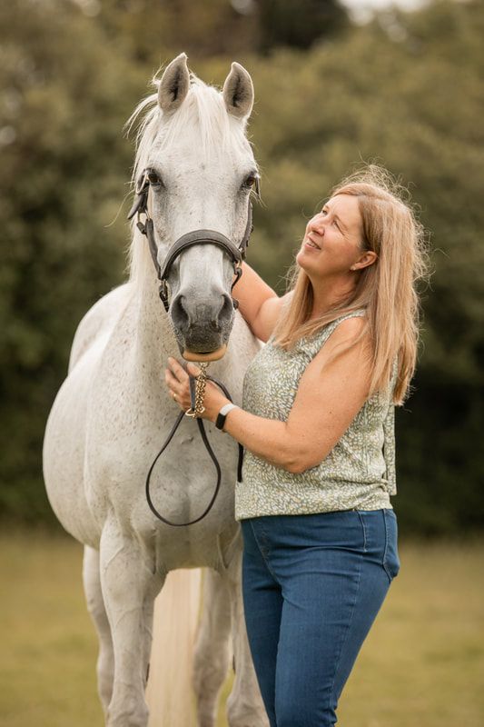 A beautiful white horse stands in a field while being lovingly stroked by someone in a floral top and jeans.