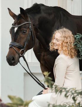 A bride in a white dress shares a tender moment with a dark horse against a white wall background.
