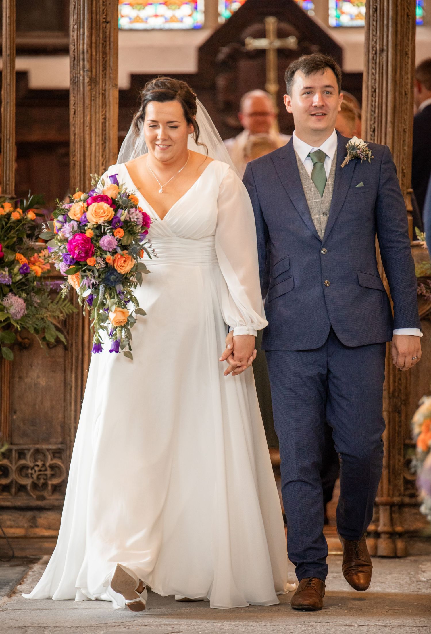 A couple in wedding attire stands together outdoors, with the bride in white and groom in navy suit.