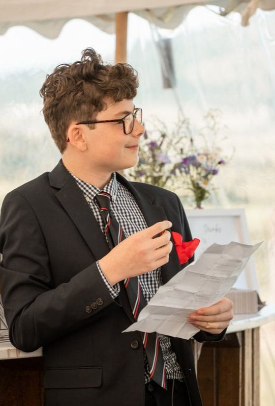 A person in a dark suit and checkered shirt reads from papers while holding a red boutonniere at a formal event.