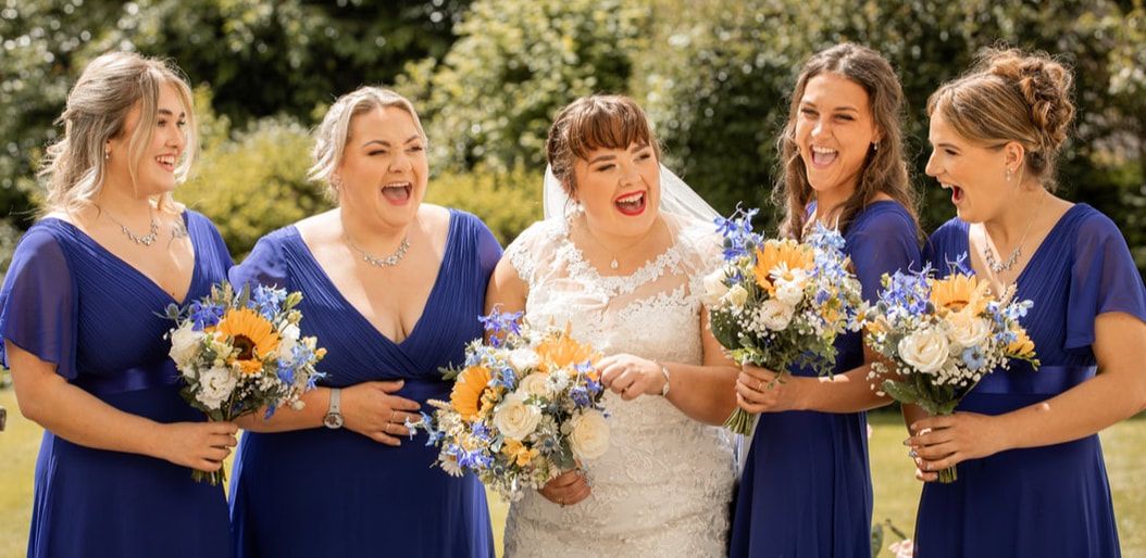 Wedding party members in navy blue dresses and white gowns share joyful moments while holding floral bouquets outdoors.